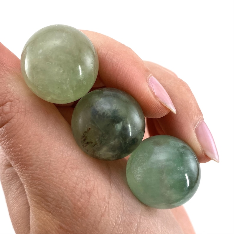 May include: Three green fluorite spheres, each with a slightly different shade of green and white, are held in a hand. The spheres are smooth and polished.