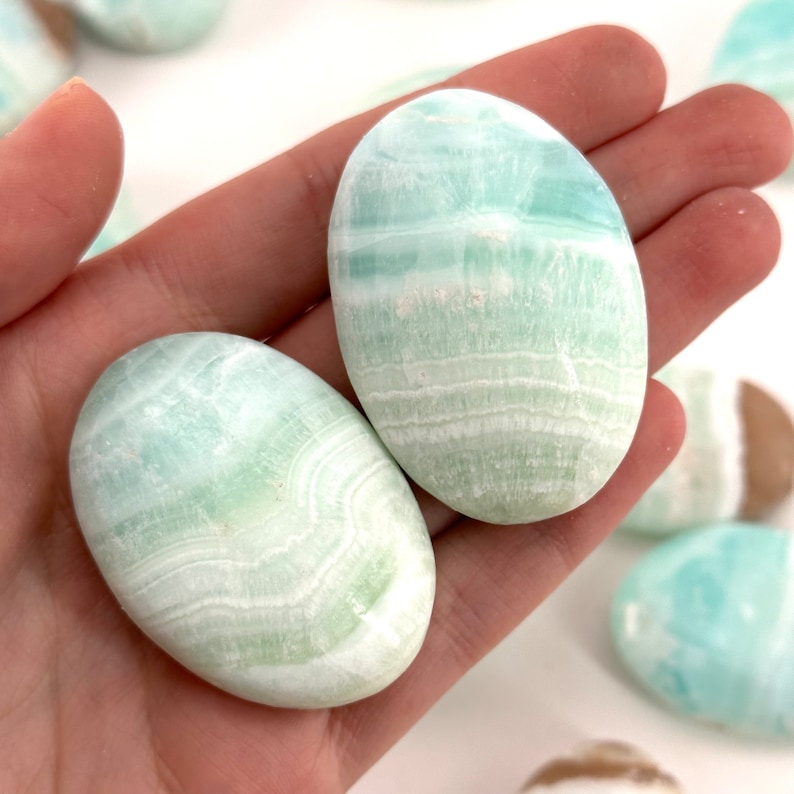 Op de afbeelding: Twee gladde, ovale groene en witte celestiet palmstenen. De stenen hebben een gebandeerd patroon en zijn gepolijst.
