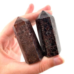 May include: Two dark red and brown gemstone points, also known as red garnet, are held in a hand. The points are polished and have a smooth, shiny surface.