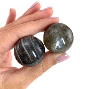 May include: Two black and grey striped crystal spheres, also known as obsidian spheres, are held in a hand. The spheres are smooth and polished.