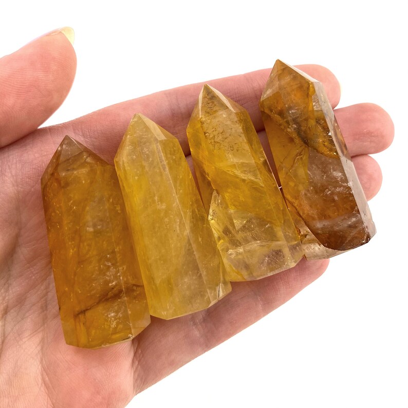 May include: Four golden-yellow quartz crystal points are held in a hand. The crystals have a translucent appearance with variations in color, ranging from light yellow to a deeper amber hue. The crystals are polished and have a faceted shape.