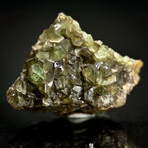 May include: A close-up of a rough, uncut mineral specimen. The stone is a mix of olive green and brown hues, with many small, crystalline structures. The surface reflects light, creating a sparkling effect. The overall shape is irregular.