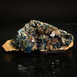 May include: A close-up of a mineral specimen featuring a mix of colors. The rock has a rough, textured surface with dark brown, blue, and tan hues. Some areas have a crystalline appearance. The mineral is displayed against a dark background.
