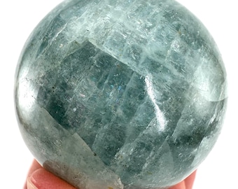 Puede incluir: Una esfera translúcida de color azul verdoso pálido con una superficie facetada. La bola de piedra preciosa se sostiene en una mano, mostrando sus cualidades pulidas y reflectantes. El color de la esfera es una mezcla de azul y verde.
