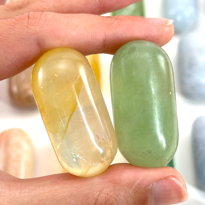 May include: Two smooth, polished stones, one yellow and one green, are held in a hand. The stones are oval shaped and have a glossy finish.