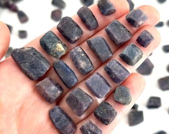 May include: A collection of raw, rough, dark purple sapphire crystals. The crystals are various sizes and shapes, with some having a slightly metallic sheen.
