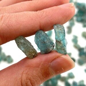 May include: Three small, rough, blue-green gemstones, likely apatite, are held in a hand. The stones are translucent and have a slightly cloudy appearance.