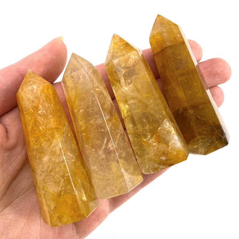May include: Four polished citrine crystal points in varying shades of yellow and gold are held in a hand. The crystals have a faceted, tapered shape, and are displayed against a white background.