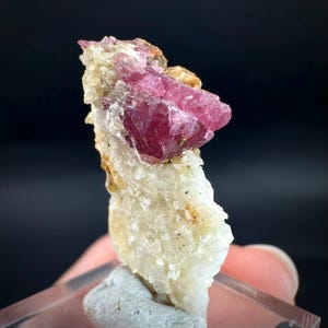 May include: A striking pink tourmaline crystal formation set on a base of off-white quartz. The tourmaline exhibits a deep, vivid colour and a crystalline structure. The quartz base has a rough, textured appearance, with small flecks of other minerals.