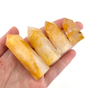 May include: Four golden yellow quartz crystal points with varying shades of yellow and white. The crystals are held in a hand, showcasing their polished, pointed tips and natural patterns. The crystals are a natural stone.