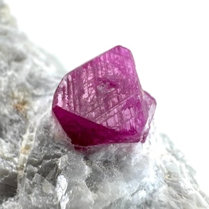 May include: A close-up of a vibrant, faceted ruby crystal, showcasing a deep pink hue. The gemstone is set against a backdrop of a light gray, textured mineral, highlighting its natural beauty. The ruby's surface reflects light, revealing intricate internal patterns.