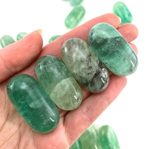 May include: A collection of green fluorite tumbled stones. The stones are smooth and polished, with a variety of shades of green. They are arranged in a row, with the largest stone at the top and the smallest stone at the bottom.