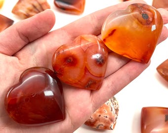 May include: A hand holding three polished heart-shaped carnelian stones, with other stones in the background. The stones range in color from deep red to orange, with natural patterns and variations. The stones are smooth and reflective.