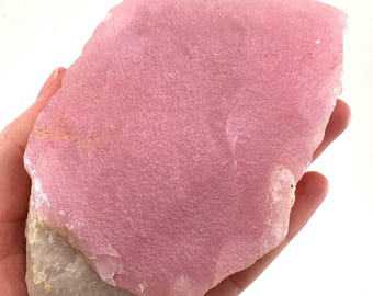 May include: A large, rough-cut piece of pink rhodonite gemstone. The stone has a textured surface and a pale pink color, with a white base. The gemstone is held in a hand against a white background.