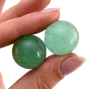 May include: Two green fluorite spheres, one with a lighter green hue and the other with a darker green hue, are held in a hand.