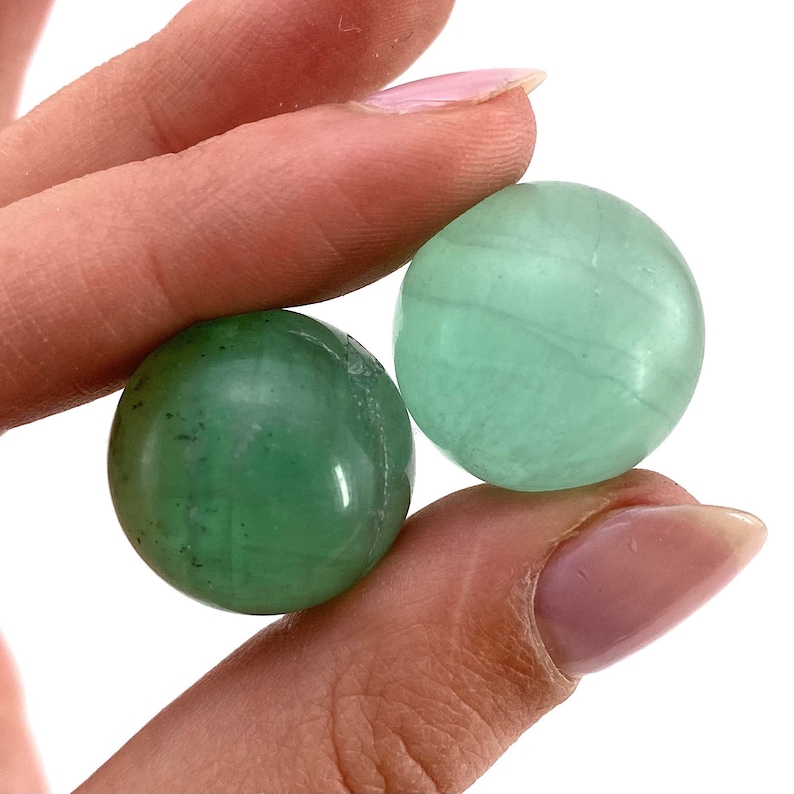 May include: Two green fluorite spheres, one with a lighter green hue and the other with a darker green hue, are held in a hand.