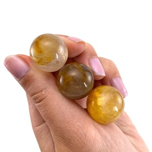 K&ouml;nnte beinhalten: Drei goldene Citrin-Kristallkugeln, jede mit einem anderen Farbton von Gelb und Braun, werden in einer Hand gehalten.