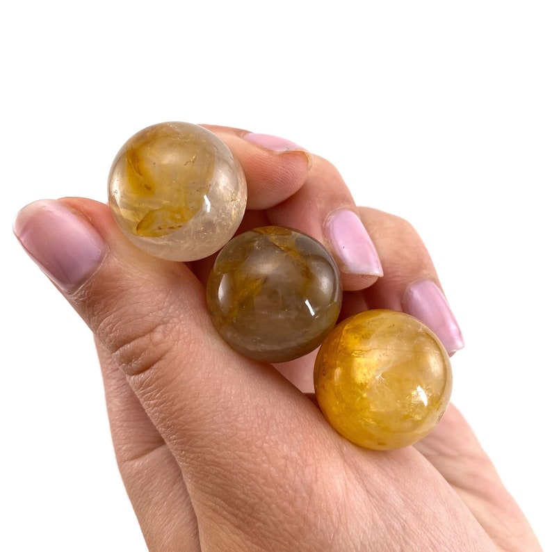 K&ouml;nnte beinhalten: Drei goldene Citrin-Kristallkugeln, jede mit einem anderen Farbton von Gelb und Braun, werden in einer Hand gehalten.