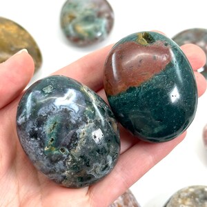 May include: Polished gemstone stones in shades of green, red, and grey. The stones have a smooth, rounded shape and are held in a hand. The gemstones are a natural product.