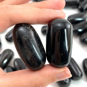 Op de afbeelding: Twee gladde, zwarte obsidiaan-tumblestones, elk ovaalvormig, worden in een hand gehouden. De stenen zijn gepolijst en hebben een glanzende afwerking.