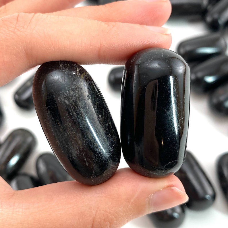 Op de afbeelding: Twee gladde, zwarte obsidiaan-tumblestones, elk ovaalvormig, worden in een hand gehouden. De stenen zijn gepolijst en hebben een glanzende afwerking.