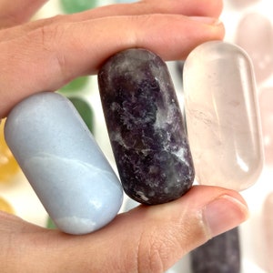 May include: Three tumbled stones, one light blue, one purple, and one clear, are held in a hand. The stones are smooth and polished.