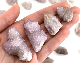 May include: Several small, light purple quartz crystal clusters are held in a hand. The crystals have a textured, sparkling appearance with hints of gold and are surrounded by other smaller, similar crystals. The background is white.