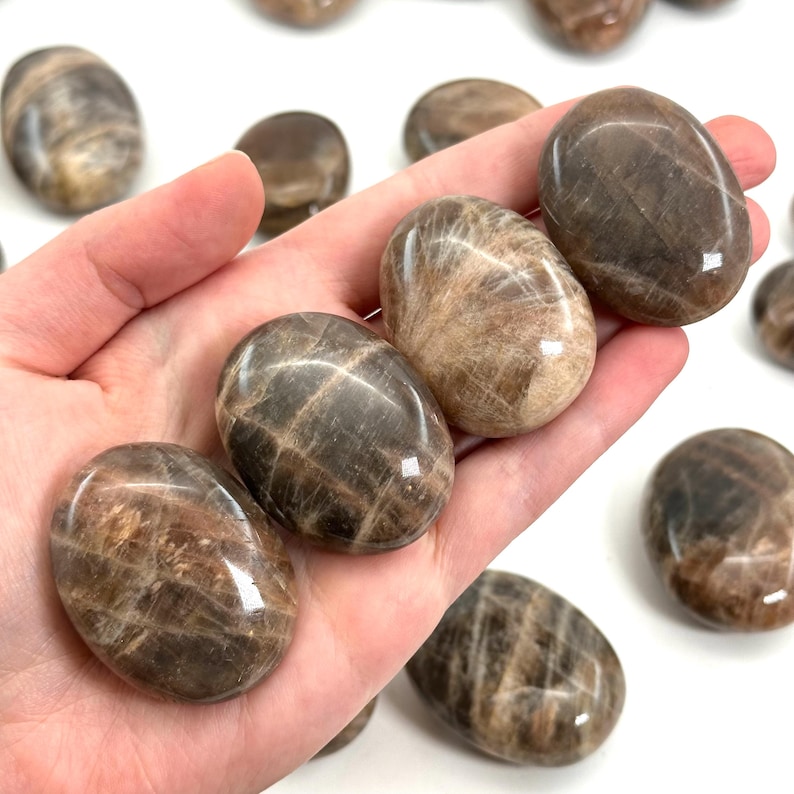 May include: A hand holding several polished, oval-shaped stones. The stones have a mix of brown and tan hues with a glossy finish. Other stones are scattered in the background, creating a collection of natural, earthy gemstones.