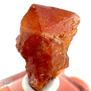 May include: A deep red, translucent, and faceted crystal with a pointed top. The crystal is on a white background.