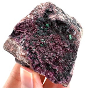 May include: A close-up of a raw mineral specimen. The rock displays a mix of colours, including light grey and tan, with patches of deep purple, black, and small green spots. The texture appears rough and crystalline.