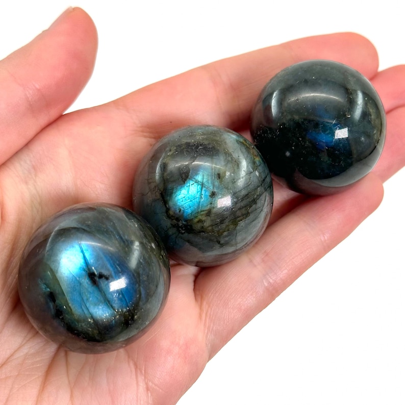 May include: Three polished labradorite spheres, each displaying iridescent flashes of blue and green. The spheres are held in a hand, showcasing their smooth, rounded forms and natural patterns. The stones have a dark, semi-translucent appearance.