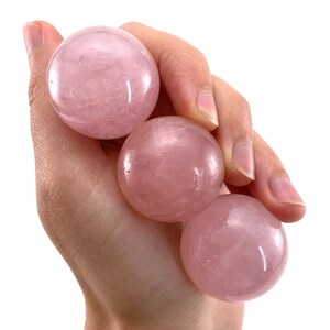 May include: Four smooth, pink rose quartz spheres, each with a slightly different shade of pink, are held in a hand. The spheres are polished and have a translucent appearance.