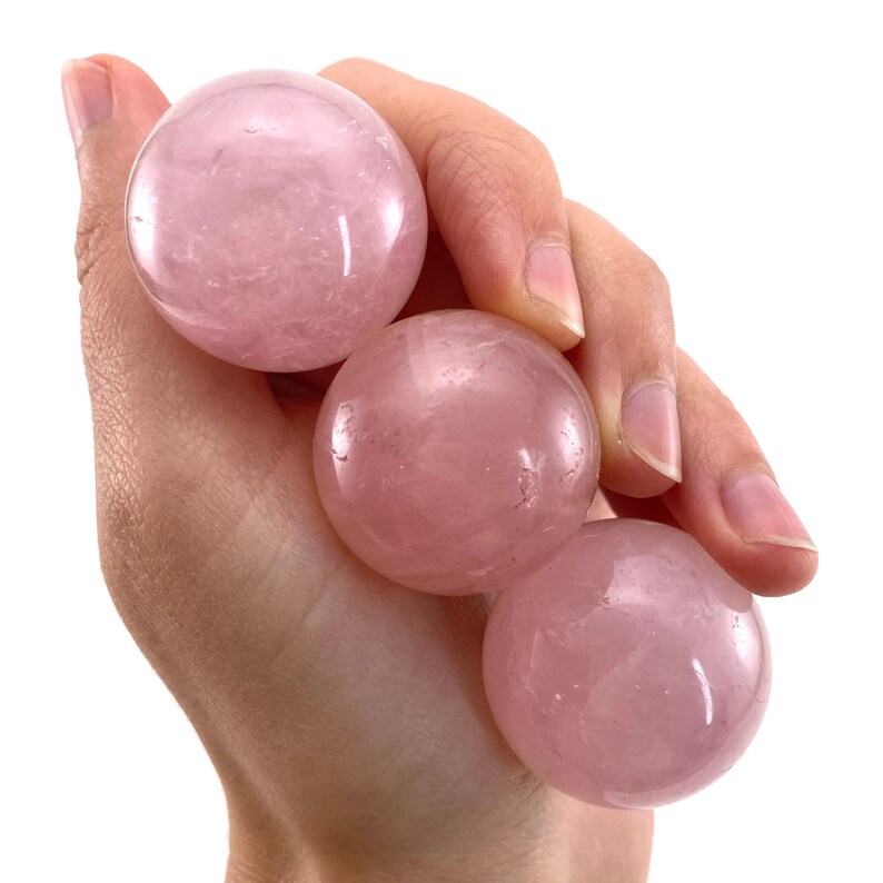 May include: Four smooth, pink rose quartz spheres, each with a slightly different shade of pink, are held in a hand. The spheres are polished and have a translucent appearance.