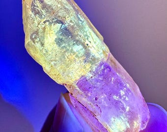 May include: A translucent, elongated crystal with a yellow-green hue at the top and a purple base. The crystal has a rough, textured surface and appears to be naturally formed. The lighting creates a gradient effect, highlighting the crystal's facets.