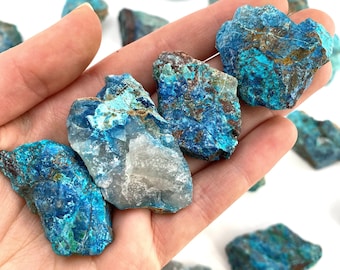 May include: A collection of rough, raw turquoise stones in various shades of blue and green. The stones have a natural, unpolished finish and are arranged in a hand.