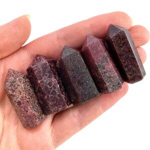 May include: Six dark red and brown gemstone points, also known as ruby in zoisite, are arranged in a row in a hand. The points are polished and have a smooth, glassy finish.
