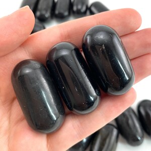 Op de afbeelding: Drie gladde, zwarte obsidiaan-tumbled stenen, elk in de vorm van een ovaal, worden in een hand gehouden. De stenen zijn gepolijst en hebben een glanzende afwerking.