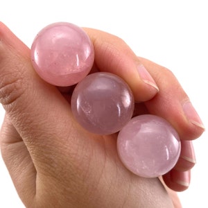 May include: Three polished rose quartz spheres in varying shades of pink are held in a hand against a white background. The spheres have a smooth, reflective surface, showcasing the natural colour variations of the stone.