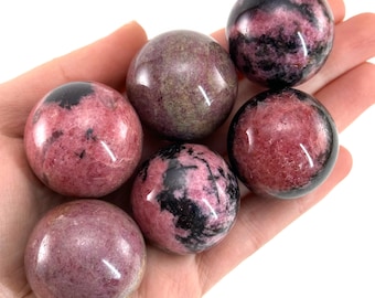 May include: Six polished gemstone spheres in shades of pink, black, and grey are held in a hand. The spheres have a smooth, glossy finish and vary in colour intensity. The gemstones are likely used for decorative or metaphysical purposes.