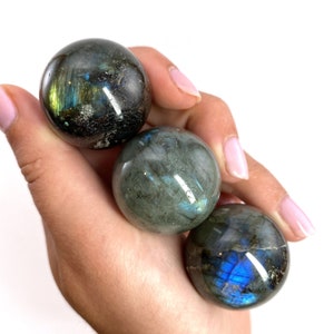 May include: Three polished labradorite spheres, each displaying iridescent flashes of blue, green, and gold. The spheres are held in a hand against a white background, showcasing the natural beauty and unique patterns of the gemstones.