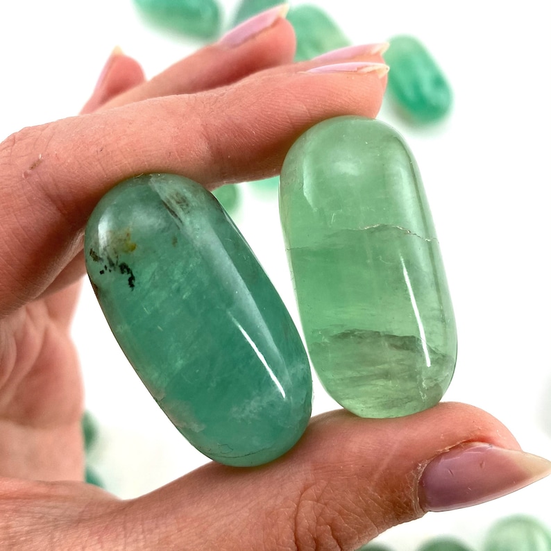 May include: Two smooth, green fluorite tumbled stones. The stones are oval shaped and have a translucent, glassy appearance.