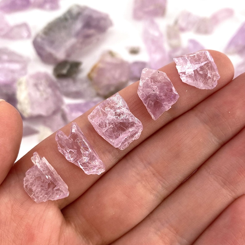 K&ouml;nnte beinhalten: Sechs rosa, durchscheinende, grob geschliffene Kristalle in verschiedenen Gr&ouml;&szlig;en. Die Kristalle sind in einer Reihe auf einer Hand angeordnet.