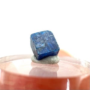 Puede incluir: Un espécimen mineral de color azul oscuro, en forma de cubo, posiblemente pirita, descansa sobre una pequeña piedra gris. El mineral tiene una superficie texturizada y se encuentra sobre un fondo borroso de color rosa claro. La presentación general es limpia y enfocada.