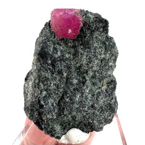 Puede incluir: Una roca rugosa de color gris oscuro con un vibrante cristal de rubí rojo-rosado facetado que sobresale de la parte superior. El rubí tiene una forma ligeramente irregular y una superficie brillante. La roca tiene una apariencia texturizada y granular.