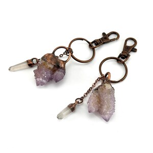 May include: Two keychains with copper-colored hardware. Each keychain features a cluster of light purple amethyst crystals and a clear quartz crystal pendant. The keychains have a lobster clasp and a split ring.