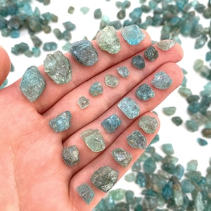 May include: A collection of small, rough, green and blue aquamarine crystals. The crystals are various sizes and shapes, and are scattered on a white surface.