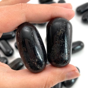 Op de afbeelding: Twee gladde, zwarte, getrommelde stenen, waarschijnlijk obsidiaan of zwarte toermalijn, worden in een hand gehouden. De stenen zijn ovaalvormig en hebben een glanzende afwerking.