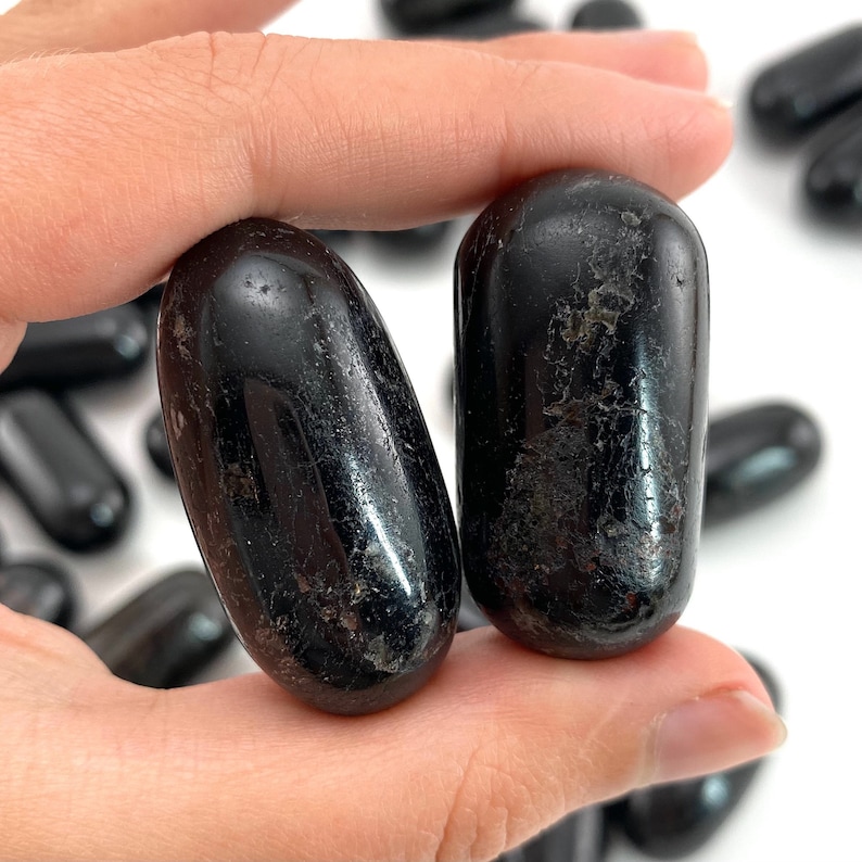 Op de afbeelding: Twee gladde, zwarte, getrommelde stenen, waarschijnlijk obsidiaan of zwarte toermalijn, worden in een hand gehouden. De stenen zijn ovaalvormig en hebben een glanzende afwerking.