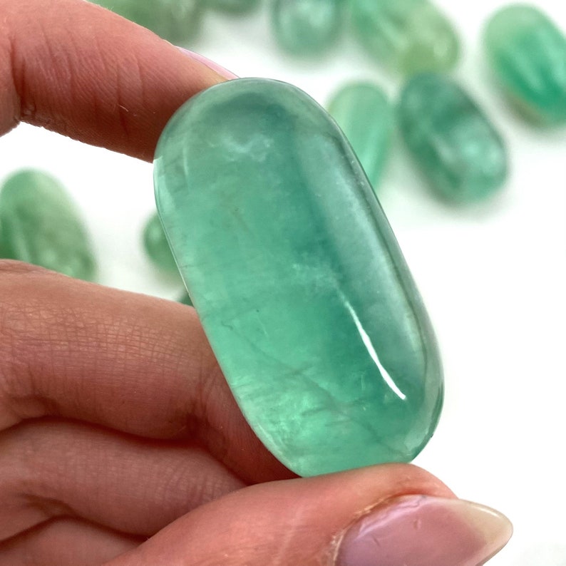 May include: A smooth, polished green fluorite stone with a light green hue. The stone is oval shaped and has a glossy finish.