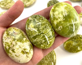 Puede incluir: Una colección de piedras de serpentina pulidas de color verde y blanco, también conocidas como ópalo verde, en varias formas y tamaños. Las piedras tienen un acabado liso y pulido y están dispuestas en una mano.